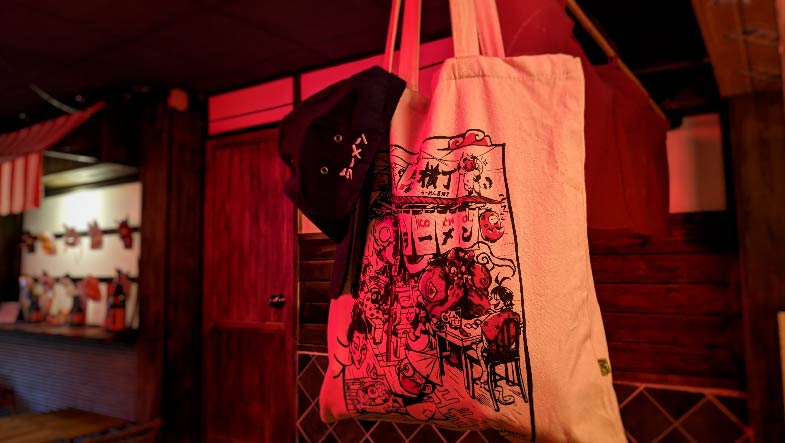 Una totebag con la ilustración de Bluwly estampada y una gorra del Yokocho colgadas en primer plano con el interior del local de Elche de fondo ligeramente desenfocado.